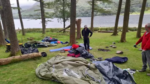 Discarded camping equipment and clothing at the side of Buttermere, with a number of chopped trees in the background