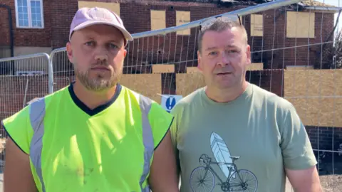 Mike has a high viz shirt on and a cap. He has a beard. Roy is stood next to him in a t-shirt and glasses on his head. He has short brown hair. They are stood in front of their damaged home which are boarded up with a metal railing surrounding them.