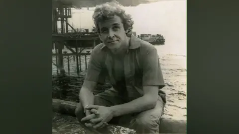 Jason Gleave Justin looks at the camera in a black and white photos. His shirt is unbuttoned and his hands are crossed. He's sitting by  the seaside with a pier in the background.