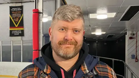 A man with short grey hair and a grey and brown beard. He stands beside an ice rink. He is wearing a black hoodie and a orange and blue checked hoodie over the top.