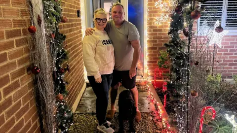 Becky and Andy Lauder at their house in Milden Gardens, Frimley Green. They are stood in front of their front door with their black dog. A number of candy canes can be seen dotted along the garden and a decoration with lights and baubles is framing the door.