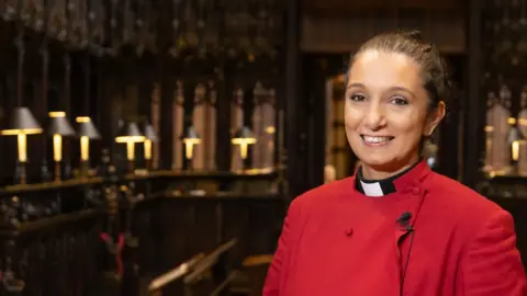 Manchester Cathedral Canon Grace Thomas with black hair tied back and wearing a read cassock and black and white clerical collar standing inside Manchester Cathedral. She is smiling.
