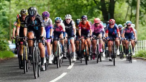 A group of cyclists in colourful racing kits ride closely together on a tree-lined road during a road race, with several riders competing for the lead.
