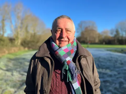 A headshot of an older man in a burgundy jumper and brightly coloured checkered scarf. He is smiling into the camera and the sky is bright blue in the background. He is bald and slightly squinting as the sun is in his eyes. He is wearing a khaki jacket and standing in a field. 