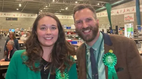Laura Coffey/BBC A woman in a green top stands next to a man in a green shirt and brown blazer.