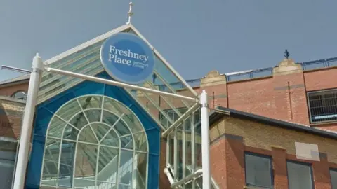 The glass canopied Entrance to Freshney Place with a blue name sign above the door and the red brick multi-storey car park in the background