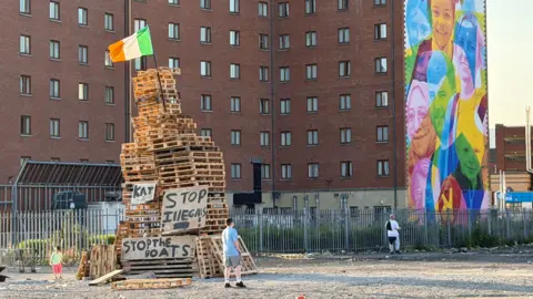 An Irish tri-colour is on top of a small, unlit bonfire. Signs saying 'STOP ILLEGALS' and 'KAT' are placed upon it too. Around it is wasteground. 