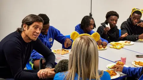 Neil Sherwood/Children In Need/PA Wire BBC broadcaster Vernon Kay interviewing a youth leader at the youth centre with long blond hair and wearing Children in Need headband while other young people watch on as they eat food  