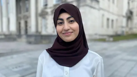 A teenage girl wearing a head covering smiles at the camera. She is stood near a white civic building in Leeds.