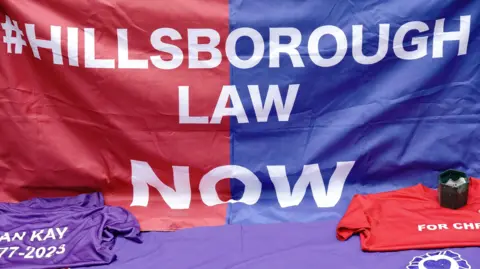 A Hillsborough Law banner. It reads #Hillsborough Law Now - blue on one half and red on the other with white lettering. Two t-shirts - one purple, the other red - are in front of it, along with a candle.