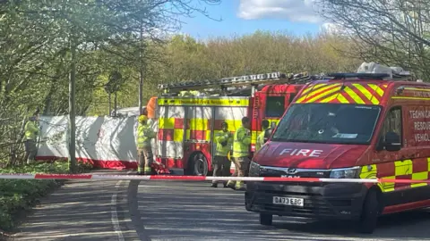 A road is cordoned off by red-and-white tape with two fire and rescue vehicles parked across the carriageway. Four firefighters are visible wearing wearing high‑visibility clothing. A tall white screen has been erected next to the road, partially blocking the view. The scene is a tree‑lined road under a bright sky.