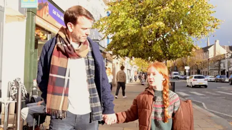 Cockermouth and District Chamber of Trade A young girl with red hair and plaits holds a man's hand as they walk down a street together in the sunshine. 