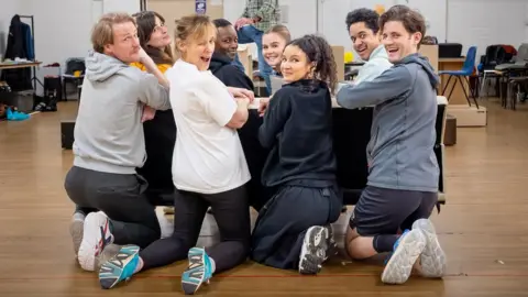 Marc Brenner A group of actors on their knees around a table turn and smile at the camera