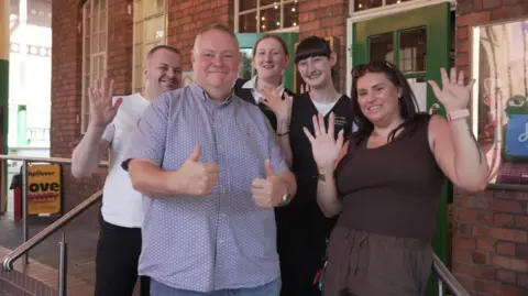 Five people are standing outside a building with their thumbs up or hands waving for the camera.