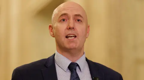 PA Media Jon speaking in the Great Hall at Parliament Buildings. He is wearing a navy suit jacket, blue and white striped top and navy tie. 