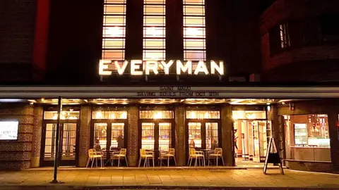 BBC The facade of an art deco-style cinema building at night. There are five patio glass doors with tables and chairs in front. The far right is the entrance to which the doors are wide open and a person can be seen walking inside. Above the doors is a lit LED sign stating: EVERYMAN.