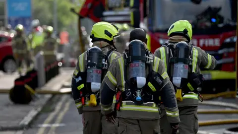 Firefighters walking down street in full gear including yellow helmets in front of fire engine with yellow hoses in front. 