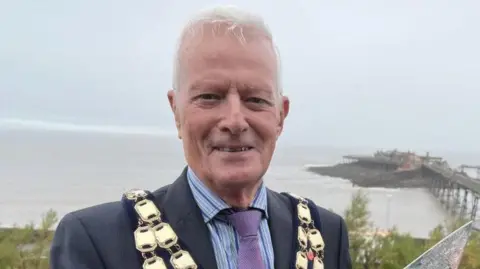 Mayor John Crockford-Hawley standing with his mayoral outfit on in a suit. In the background there is Weston pier.