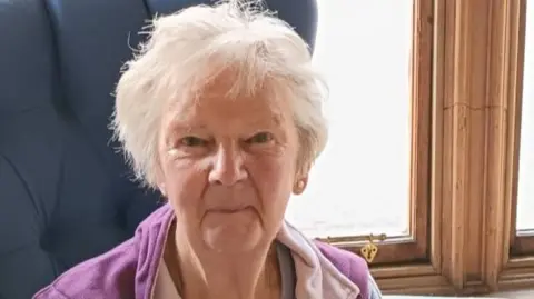 A grey haired woman with a purple top, looks directly at the camera