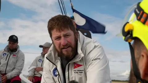 Shaun Whitmore/BBC Three crew members wearing white jackets sit on a boat. There is a blue flag flying in the background.