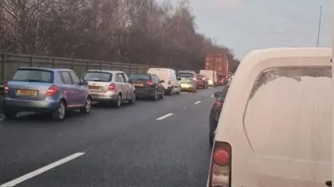 Matt Bill Queueing traffic on a dual carriageway road. A row of queuing vehicles can be seen in one lane while the back of a white van is nearest the viewer. A wooden fence and trees are beyond the queuing traffic