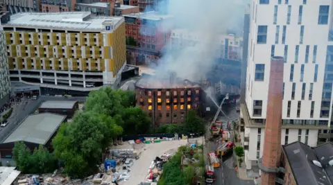JMG Press Image is an aerial shot from a drone, showing the Hotspur Press building ablaze. Fire crews on aerial ladder platforms and trying to douse the blaze. The building is next to a high-rise apartment tower block, and close to a raised railway line.