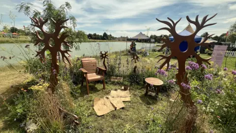 The MS garden is next to the lake at Badminton Court with many flowers of different colours and sizes. There are large wooden structures that look like nerves, with a wooden armchair and small table in the centre of a cleared space.