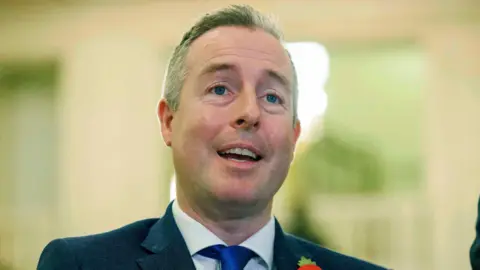 PA Media Paul Givan is wearing a white shirt, blue tie and a navy blazer. He has short grey and black hair. The top of a poppy is visible on his left hand side. The background is blurry. 