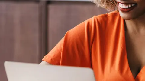 Getty Images is an interrogation image showing the lower half of the smiling woman's face looking at a laptop. She is wearing an orange top.