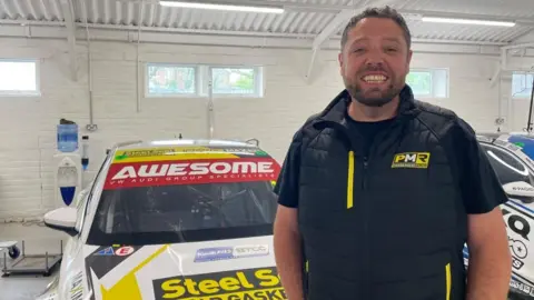 A man with short brown hair, wearing a black top with yellow PMR branding on it, stands in a garage in front of two white branded racing cars.