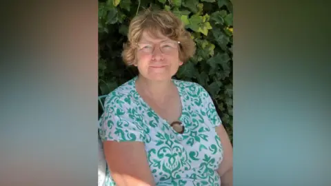 Karen Andrews A mid-shot of Karen sitting outside under greenery in a garden chair. She is smiling to camera and wearing a white and green floral summer dress