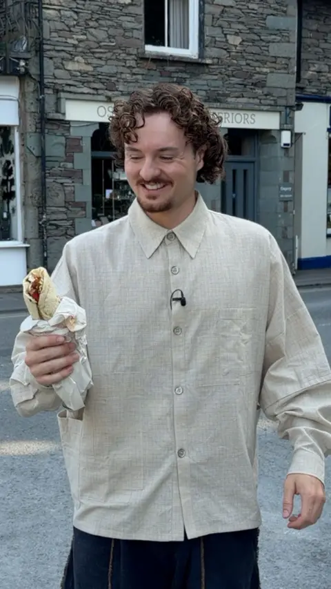 A person with curly hair and a light beige button-up shirt smiles while holding a wrapped kebab on a street in Grasmere, with traditional stone buildings in the background