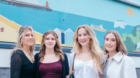 AUB/UHD Three young women with long fair hair posing in front of a large mural depicting a stylised beach and seaside in block colours with a pier, the white cliffs of Old Harry Rocks, Corfe Castle ruins, a goat, a deckchair and some beach huts.