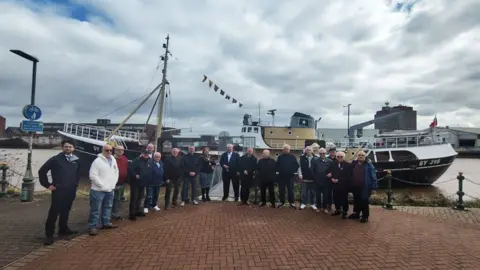 A group of people stand in front of the Ross Tiger trawler in Grimsby Docks, The ship has a black hull with white and orange superstructure and a grey funnel with a white star on it. Nautical flags fly from the main mast and the Red Ensign is on the stern staff. 