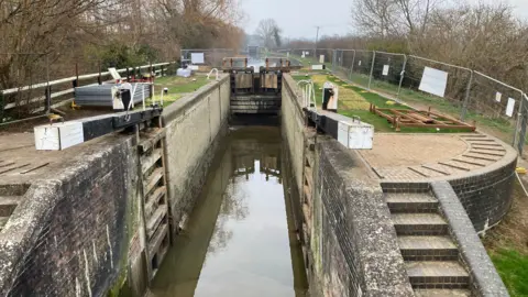 Martin Heath/BBC A lock on a canal. The gates nearest the camera are open, whilst the others are closed with a board beyond to prevent usage. There is a shallow water pool remaining in the lock, and the shadow of the lock gates is visible in the water. There is a metal fence to prevent access to the site. Stone steps from the towpath to the locks are visible to the right.