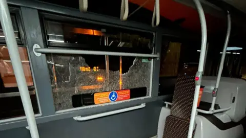 Ioan Dyer A smashed window can be seen on the bus, next to a standing area and some seats. It is dark outside.