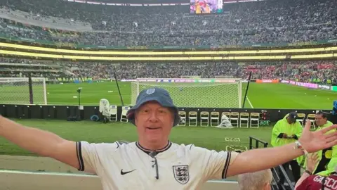 Paul Green Paul Green wears an England football shirt and a blue bucket hat. His arms are outstretched. The pitch and the stadium are behind him.