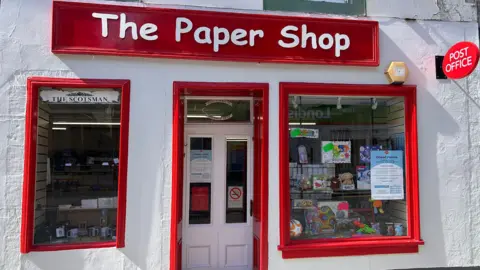 A white-walled shop with red paint trimmings and a post-office sign and its name - The Paper Shop - above the door