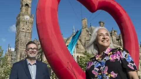 LDRS Dan Bates, wearing a blue jacket, stands to the left of the image while Shanaz Gulzar, wearing a patterned shirt, stands on the right. A red heart is between them with Bradford City Hall behind them.
