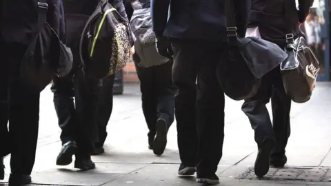 Five school pupil sets of legs, wearing black trousers and with hanging school bags, walking on stone slabs.