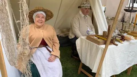 Militias of Anglia A woman and a man are sitting under a white fabric tent. The woman is wearing a straw hat with a white headscarf, and a long green and white skirt. The man is sitting behind a table, which displays items from the 17th century, including candle holders