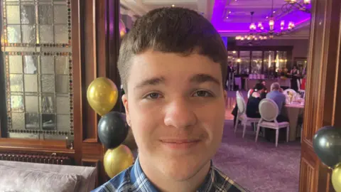 Jamie Harte is standing in an events space with different tables set out and a blue projector screen. A sofa and chairs are visible to his left. He has short brown hair and is wearing a blue chequered shirt.