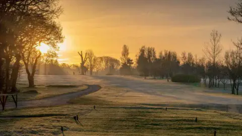A general view picture of the golf course on a pleasant morning, with the sun rising behind trees on the left of the picture. 