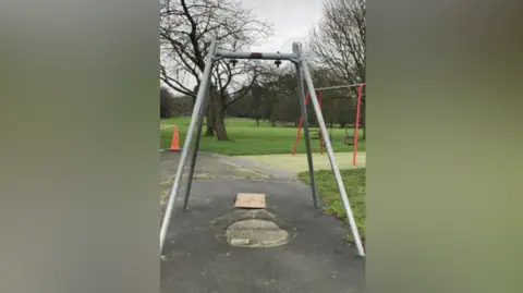 Daniel Hodgson A stand which once support a baby swing in a children's playpark. It is a large metal bracket which has worn tarmac underneath it from use. There is a red traffic cone in the background which cordons off another piece of playpark equipment. 