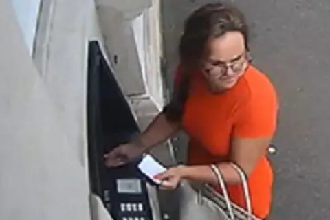 TVP Macardle attempting to withdraw money from an ATM. It looks like the card is in the machine and she's holding a piece of paper. She wears glasses and an orange dress and is carrying a bag.