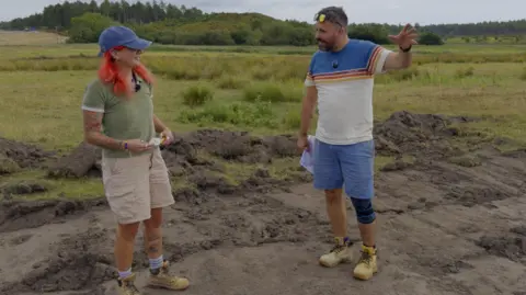 Supplied Meg Russell and Dr Derek Pitman stand at an archaeological excavation site in an open grassy landscape. Meg Russell wears excavation clothing and boots, while Dr Derek Pitman gestures during a conversation. Excavated earth and trenches are visible in the foreground, with woodland and open fields in the background.