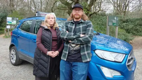 Louisa Judge Louisa Judge stands next to her son Barnaby in front of a large blue car. Louisa has long blonde hair and wears a burgundy top, black trousers and a long black coat over the top which is unzipped. Barnaby has his arms crossed in front of him. He wears a cap, glasses, chequered green and white shirt with a black T-shirt underneath and blue jeans.