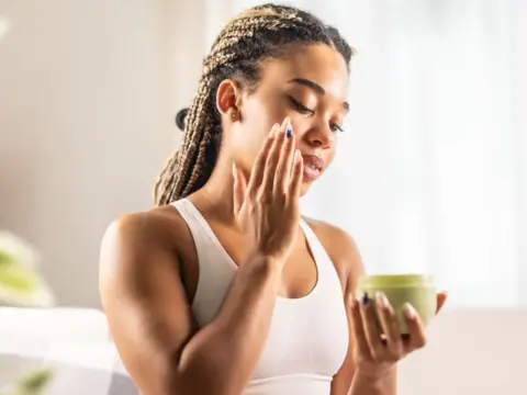 SimpleImages/Getty Images A woman with brown hair in a white top stands inside a house, applying lotion to her face from a green tub