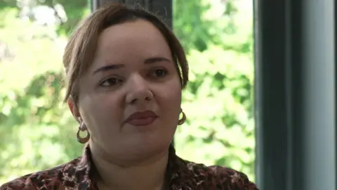 Izzy Vine has brown hair and is wearing a brown shirt with a leopard print pattern. She is sitting in a kitchen with some green trees outside the window behind her. 