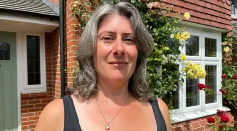 A woman with grey hair a necklace and a grey top standing in front of a house with flowers growing around the windows